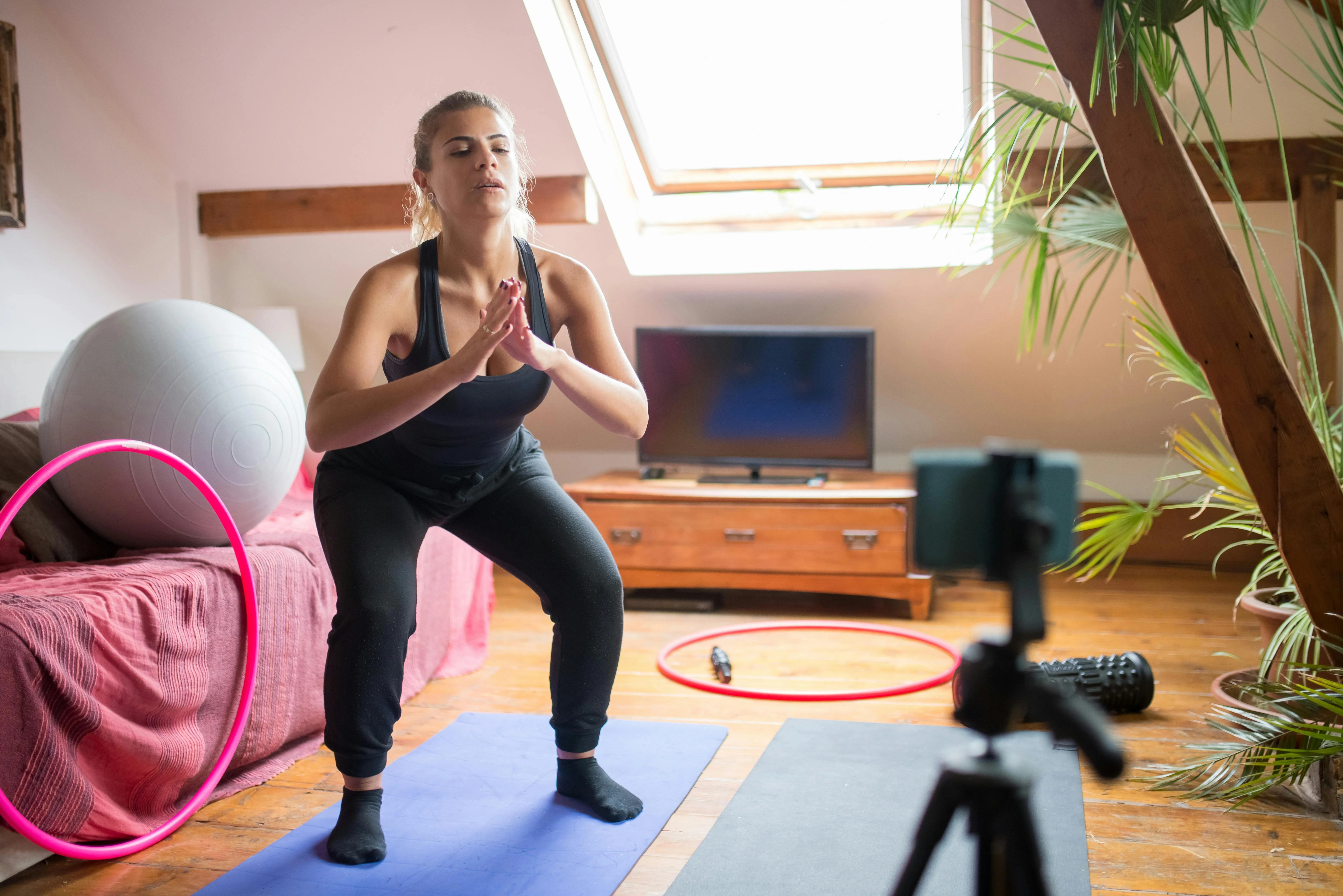 Femme en débardeur noir réalisant un squat dans son salon devant un smartphone sur trépied