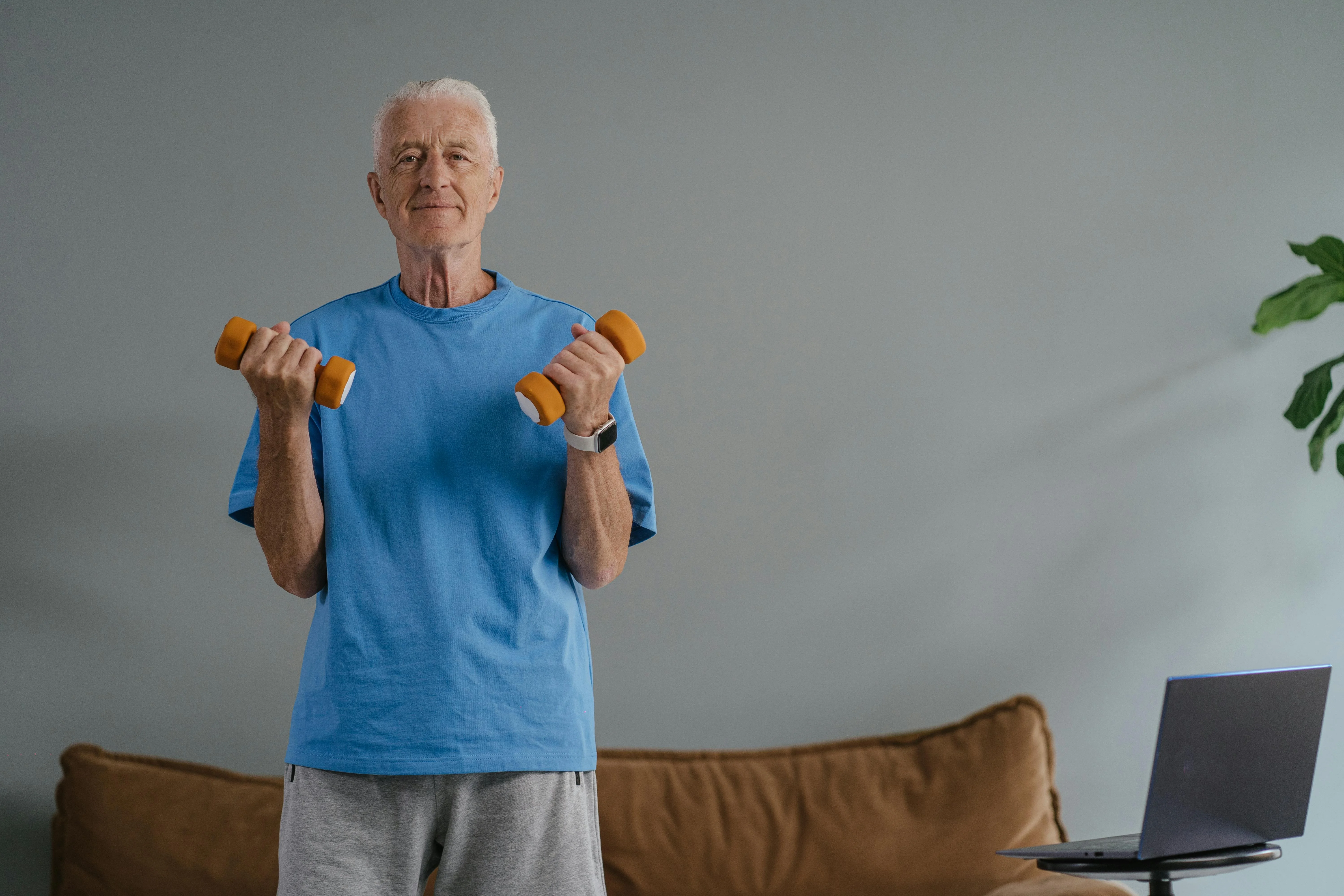 Senior souriant en t-shirt bleu fait des flexions de biceps avec deux haltères dans son salon