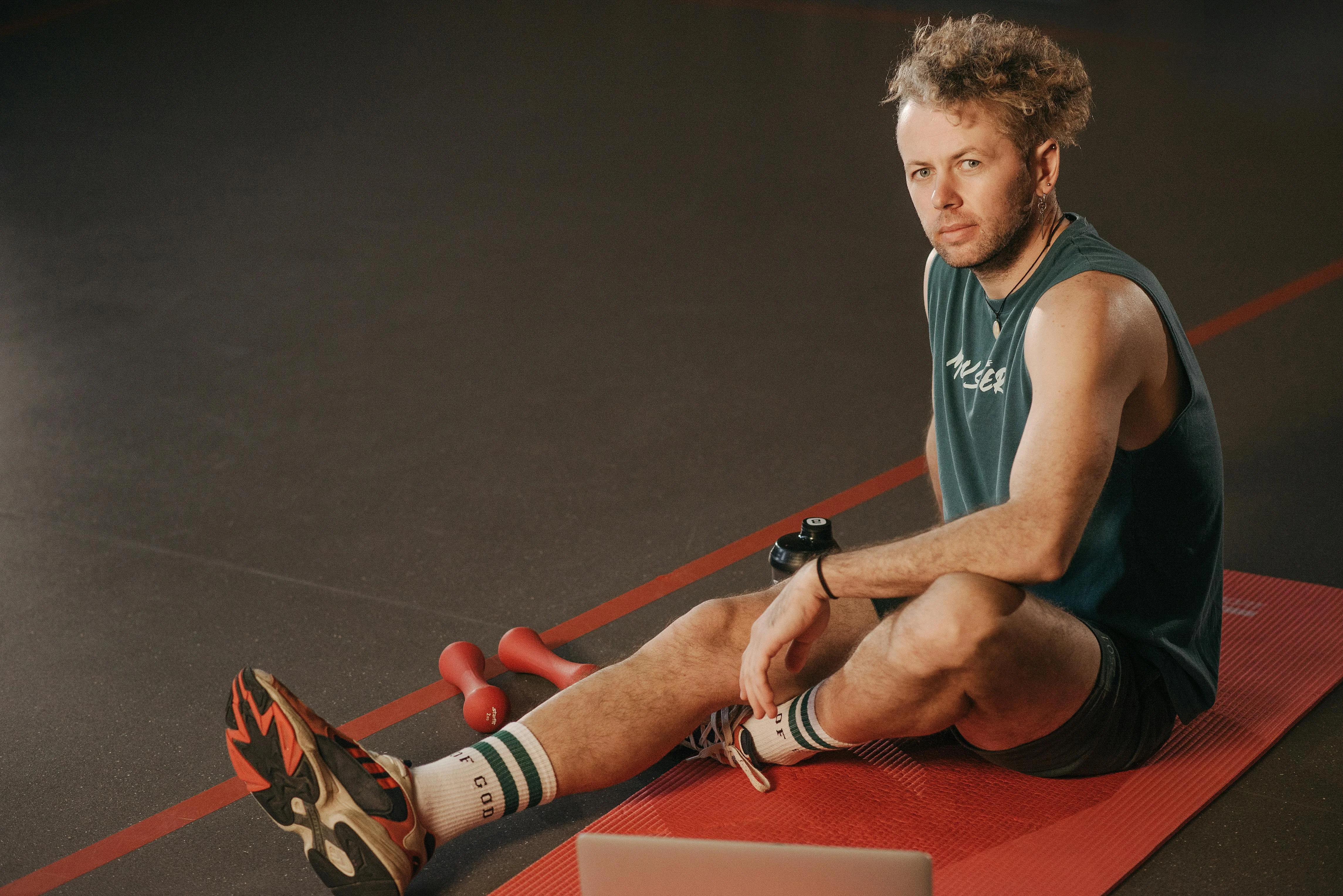 Homme en débardeur vert assis sur un tapis rouge en pause pendant une séance de reprise du sport avec petits haltères