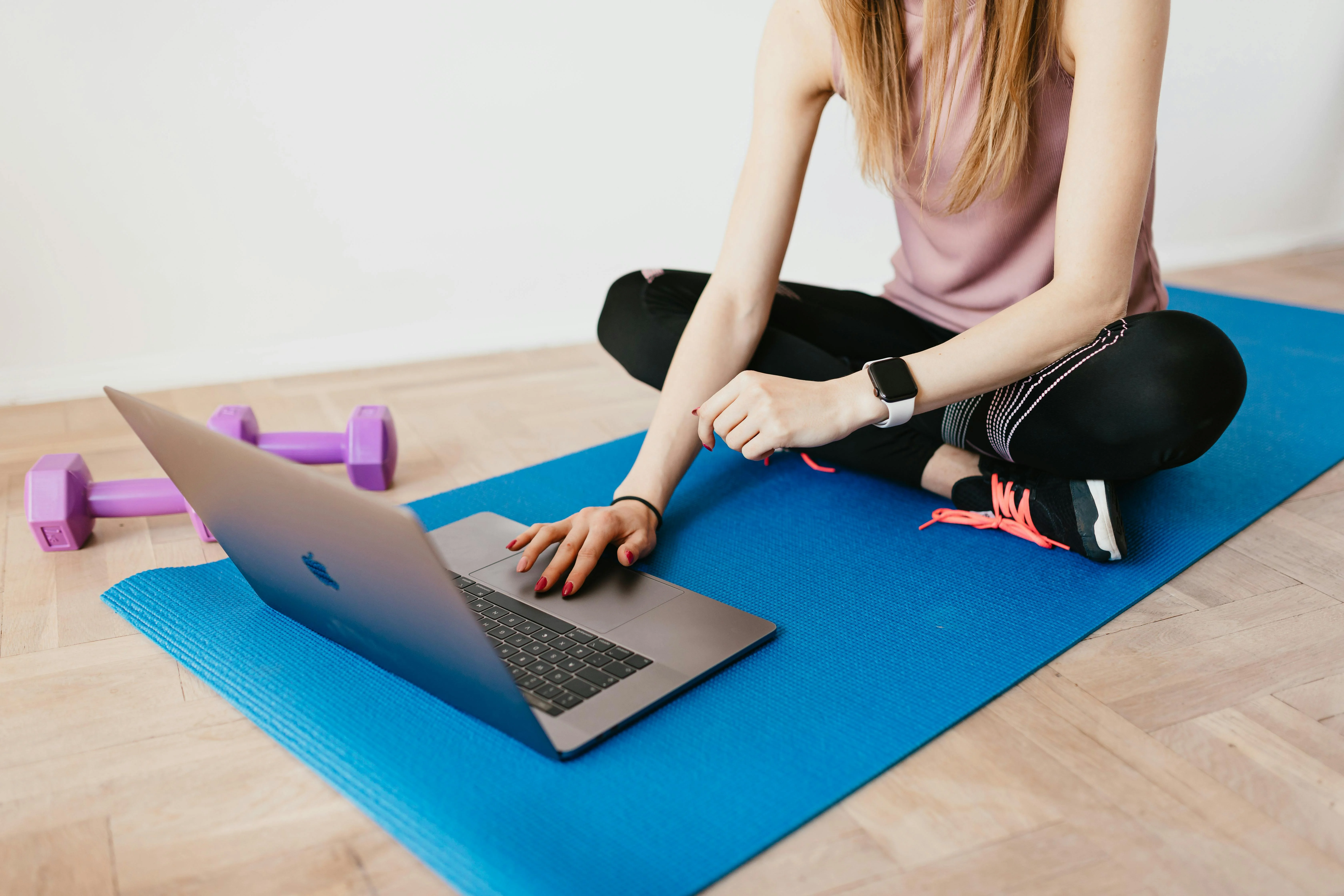 Femme en débardeur rose assise en tailleur sur un tapis bleu devant un ordinateur portable et deux haltères